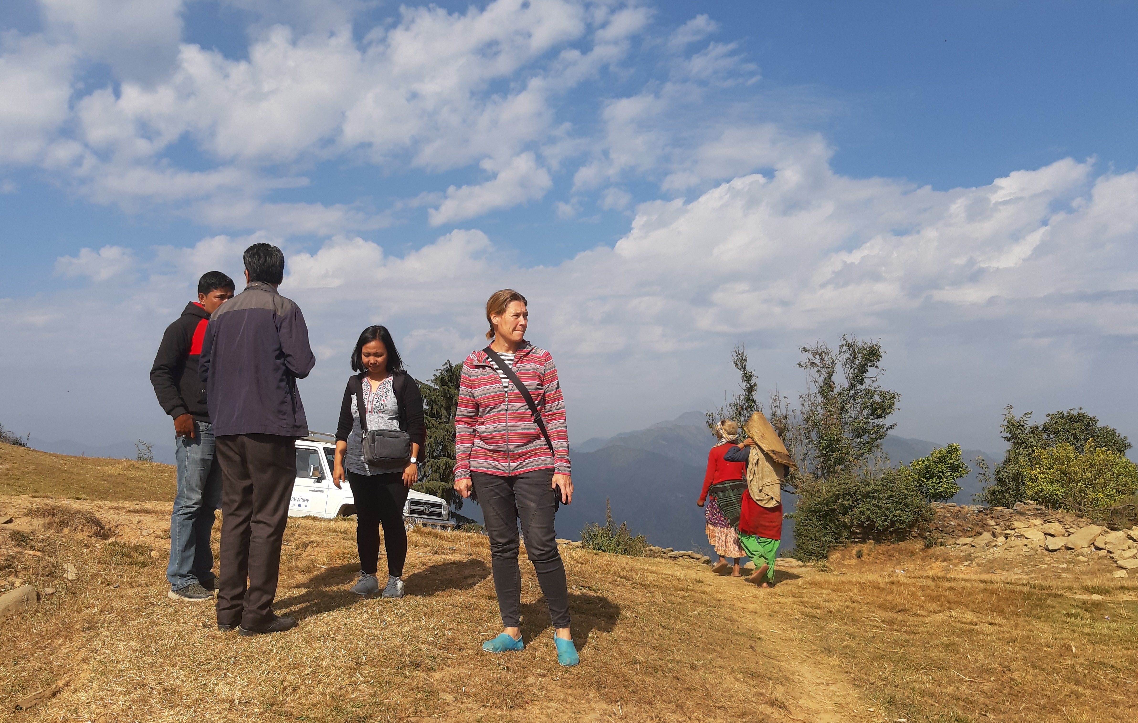Vuoren laella seisoo ryhmä ihmisiä. Taustalla kaksi nepalilaista vuoristoasukasta kulkee polkua pitkin.