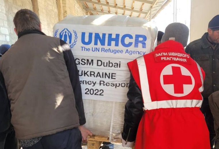 Kuvassa kaksi ihmistä kantaa lavallista Ukrainaan osoitettuja UNHCR:n avustustarvikkeita. Toisella ihmisistä on Punaisen Ristin liivi päällään.