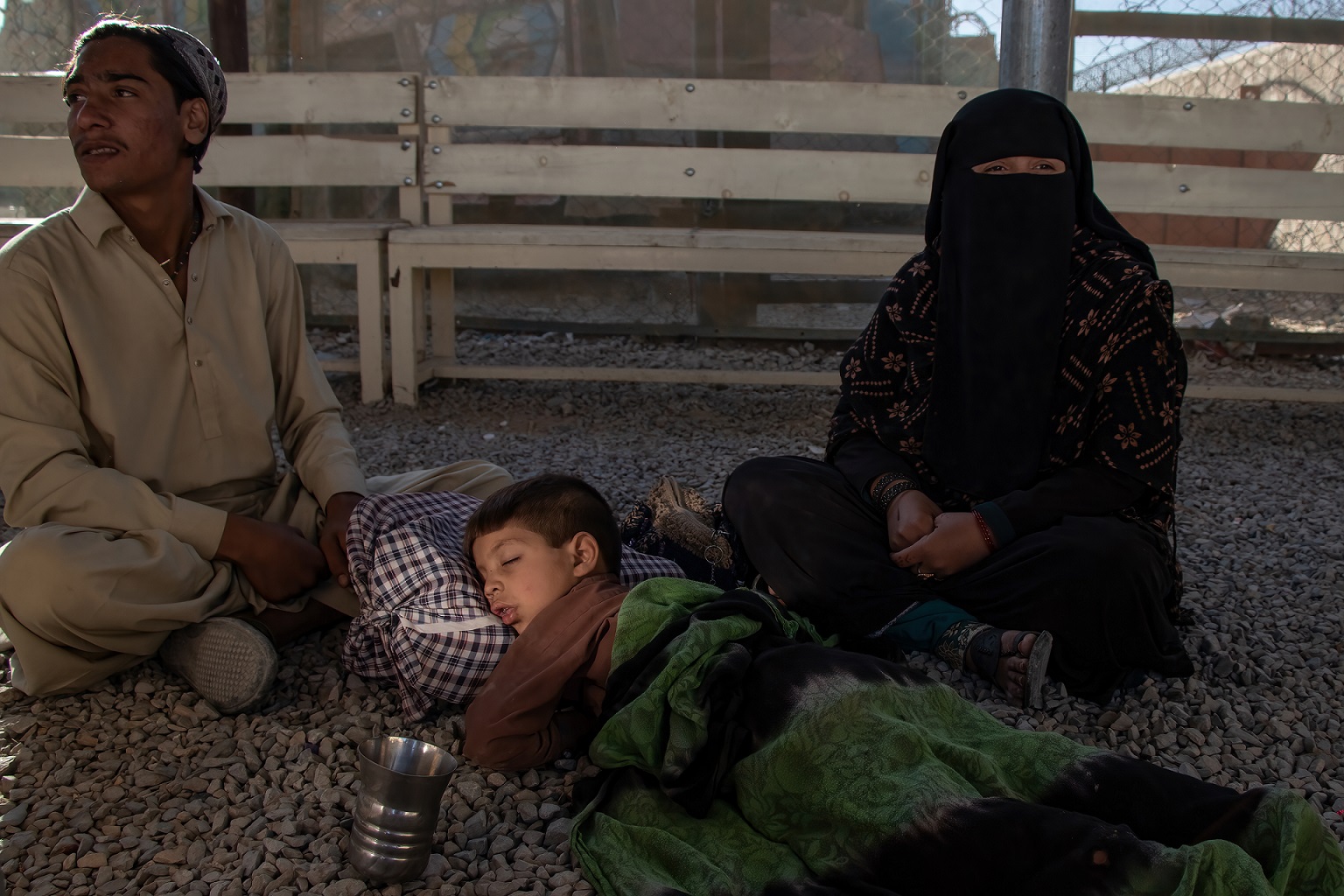 An Afghan family sitting outside. The kid is sleeping on pebbles.