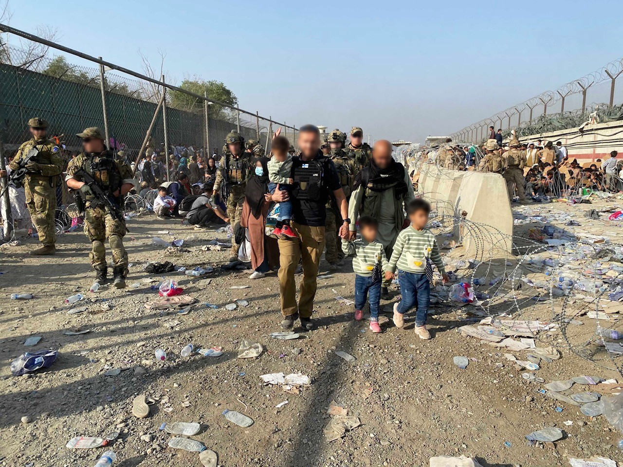 Picture of the proximity of the Kabul airfield. A member of the Finnish aid team leads two Afghan children (faces unrecognized). The picture also shows the members of the Finnish security forces and a lot of local people in the background.