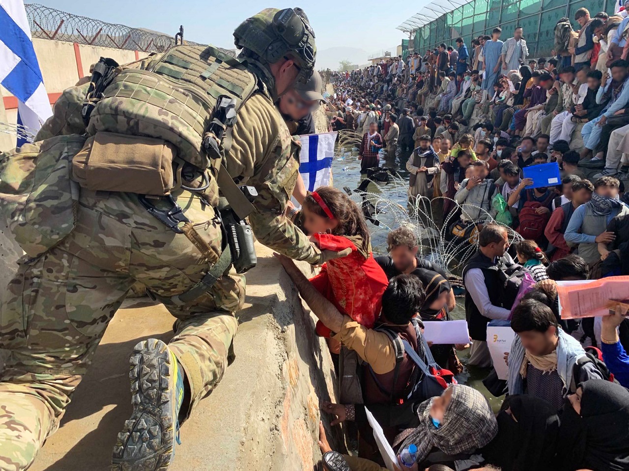 In the picture, a Finnish member of the security team helps a small Afghan girl across the wall to the shelter. In the background crowds and chaos near Kabul field.