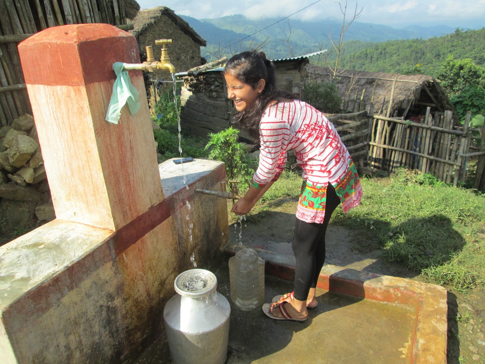Kylän vesihanoista tulee vettä astioihin. Nuori nainen on vesihanan äärellä. Vuoristomaisema taustalla