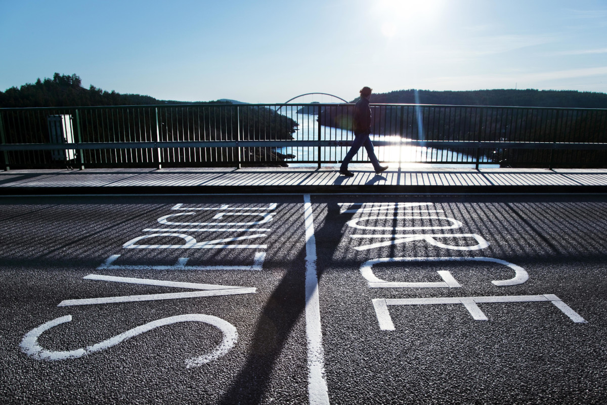 A person walks along a bridge crossing a river. Text on the bridge: Sverige-Norge.