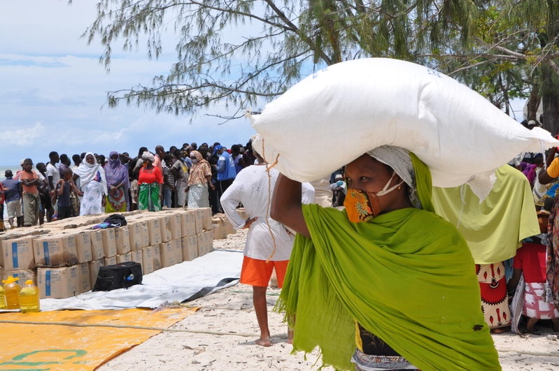Nainen kantaa säkkiä päänsä päällä. Taustalla näkyy ihmisjoukko ja avustusjärjestön laatikoita.