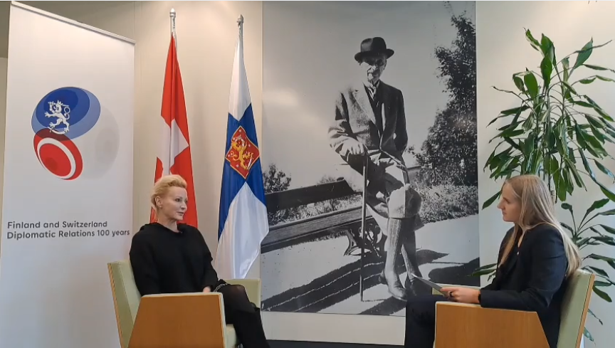 Anna Erat im Interview mit Sandra Kivimäki, beide sitzen sich in gegenüberliegenden Sesseln gegenüber. Im Hintergrund die Flaggen von Finnland und der Schweiz