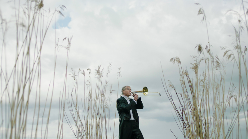 En man i frack och stående utomhus bland säv spelar trumpet.