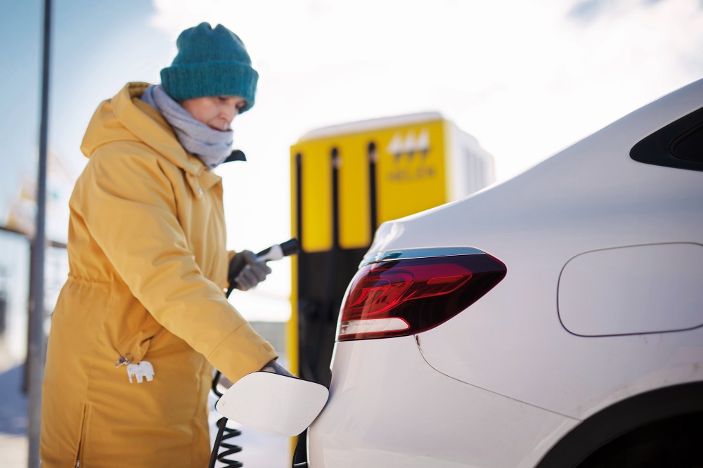 Une femme avec un manteau jaune  charge sa voiture électrique en hiver.