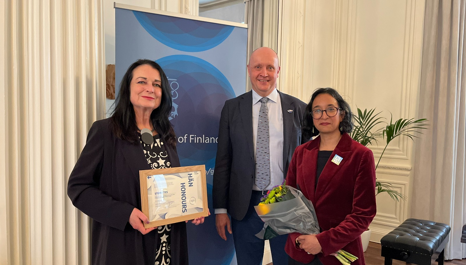 Two women and one man standing in front of a blue roll-up banner, smiling. One woman is holding flowers and the other is holding a diploma. 