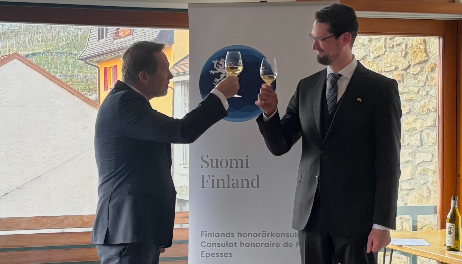 Zwei männer mit Weingläsern, im hintergrund ein Banner mit text "Finnishes Honorarkonsulat Epesses"
