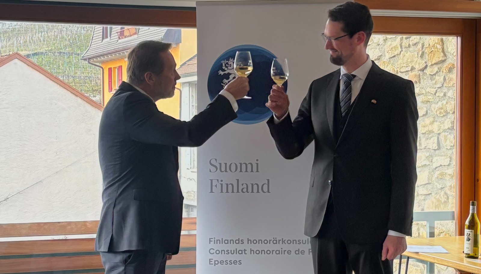 Kaksi miestä kilistelemässä viinilaseja, taustalla Suomi-banneri