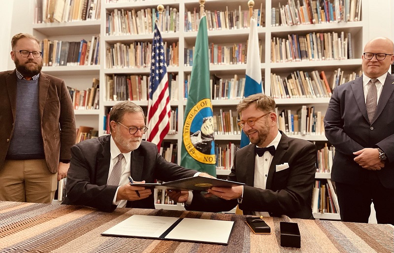 Lt. Gov. Denny Heck and Minister Harakka looking at a document, with US, FI, and WA flags and Ambassador Hautala and State Senator Marko Liias on the background.