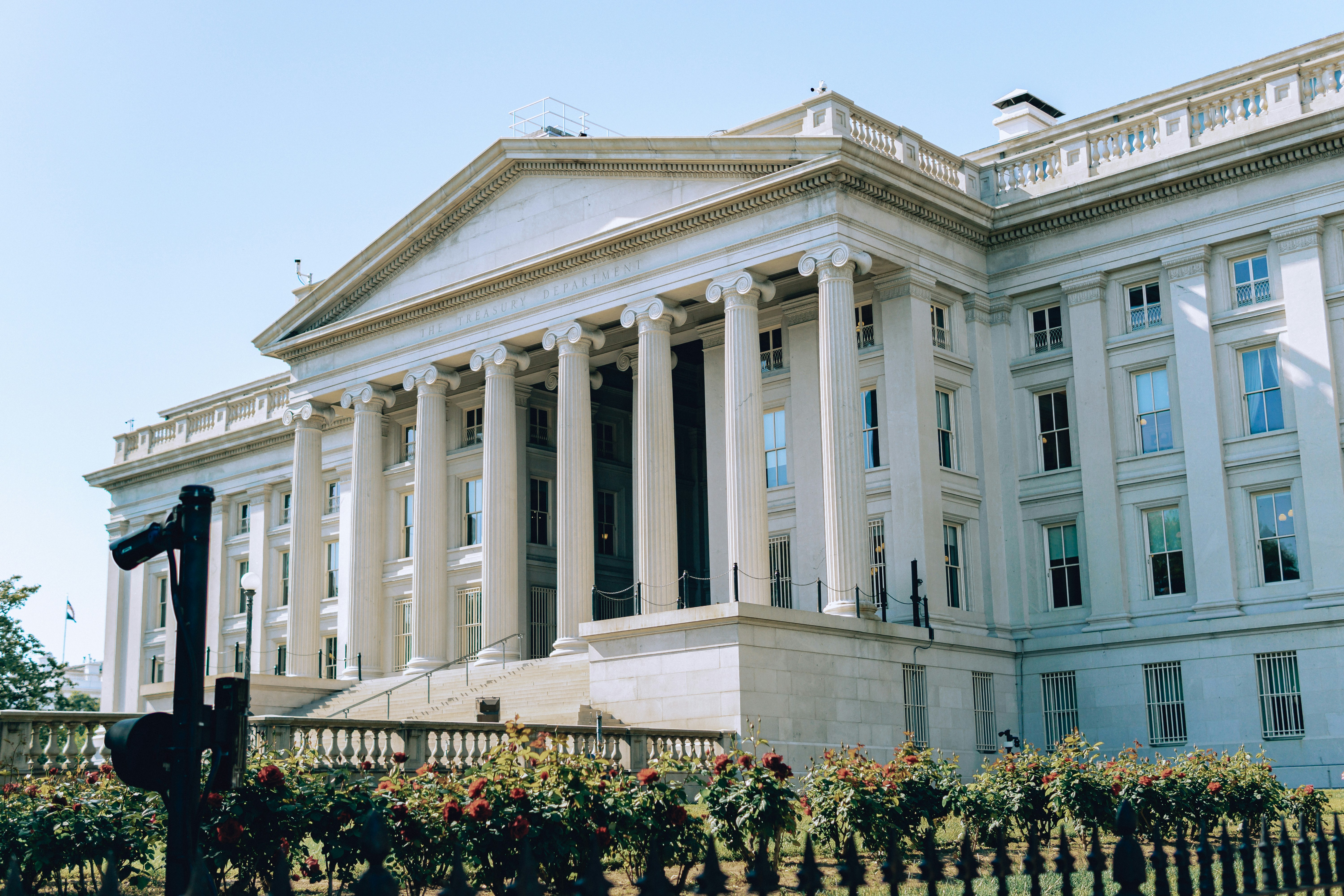 White marble building with large columns, the U.S. Treasury building, with flower bushes on the lawn.