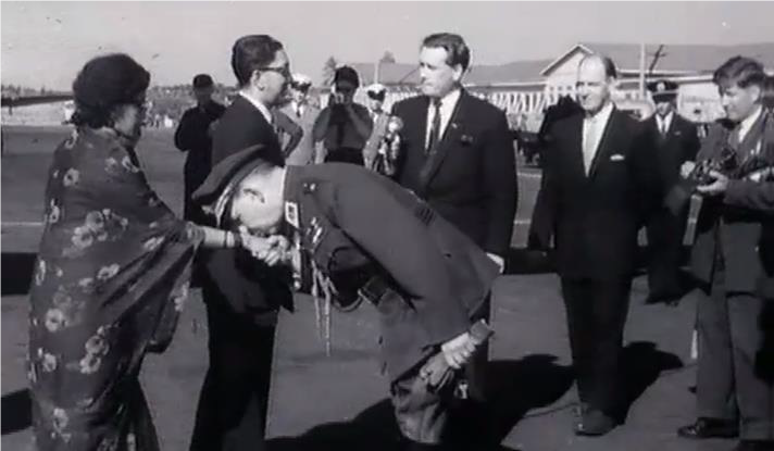 Queen and King of Nepal being greeted at the airport with adjutant in military uniform bending over to give a hand-kiss to the queen.