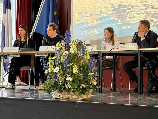 Zwei Schüler, eine Praktikantin der Vertretung und der Botschafter sitzen auf der Bühne an Tischen, vor sich Mikrofone