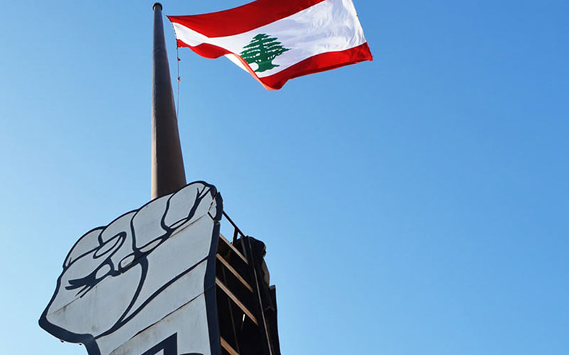 Libanonin lippu liehuu salossa. Lipun alla on nyrkkiä muistuttava kyltti. 