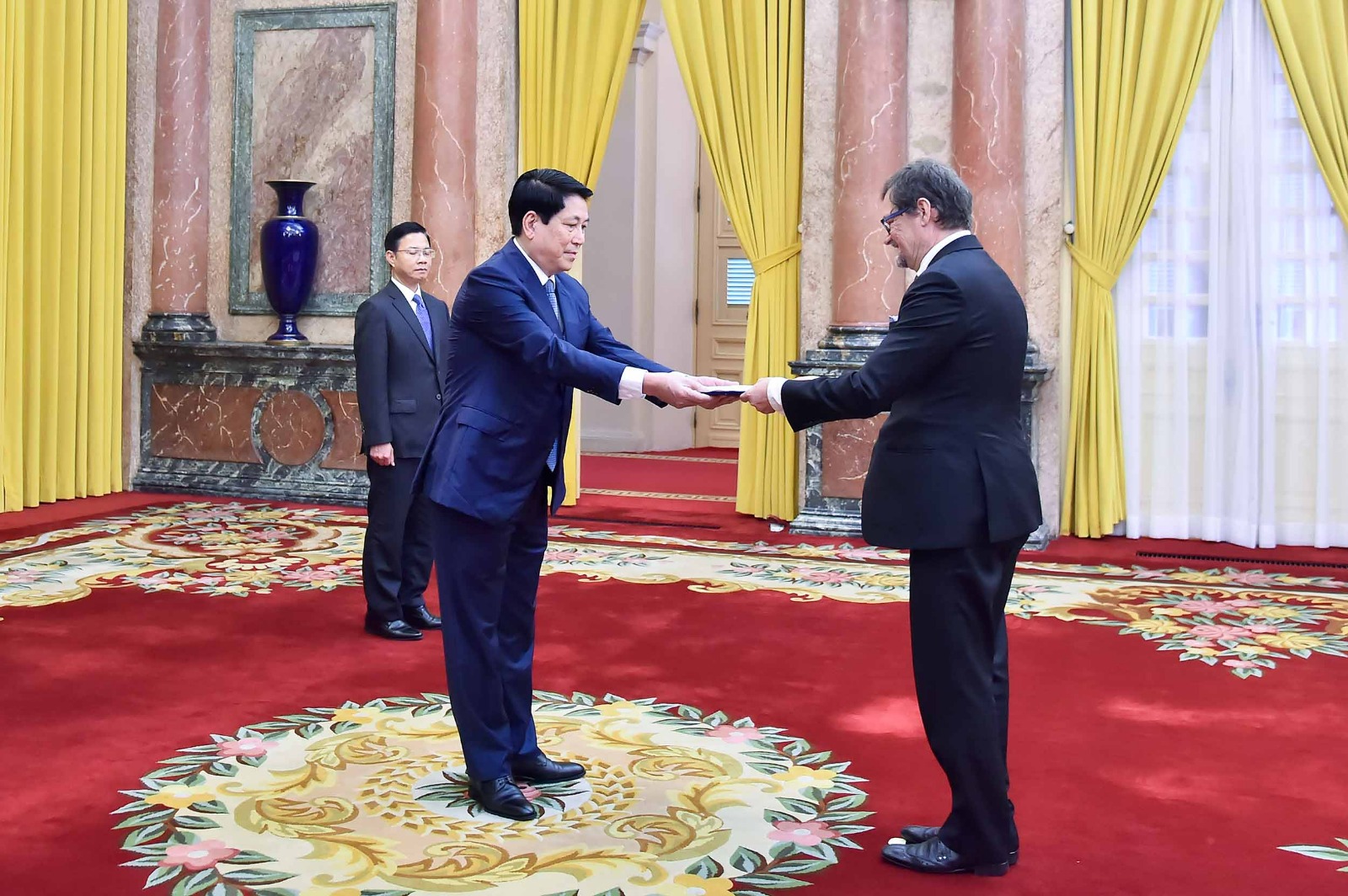 Trong ảnh có ba người, phía trước là hai người đàn ông mặc vest tối màu đangđưa một tài liệu. Phía sau là một người đàn ông khác mặc vest tối màu đang đứng.