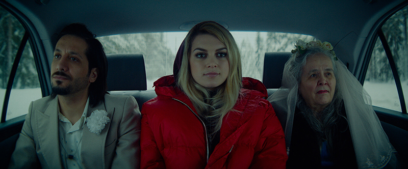 Kuva Aurora-elokuvasta: nuori nainen (päähenkilöä esittävä Mimosa Willamo) istuu auton takapenkillä juhla-asuisen miehen ja huntupäisen vanhan naisen välissä.
